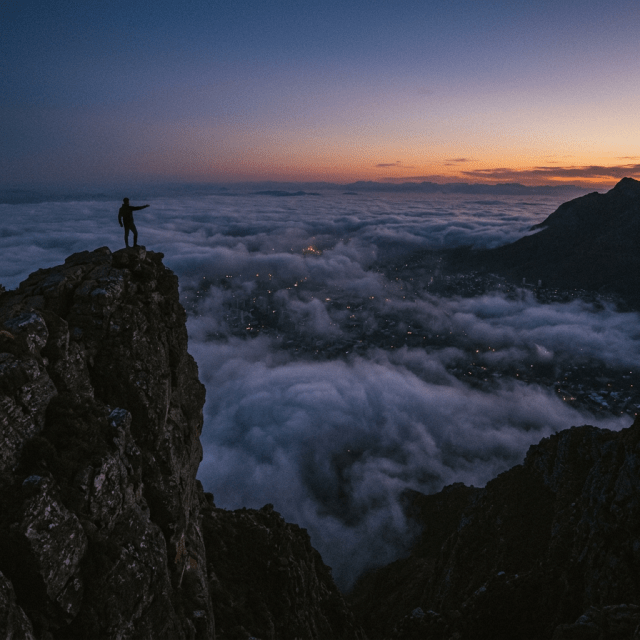 Person standing on rocky mountain peak above clouds with city lights and sunset